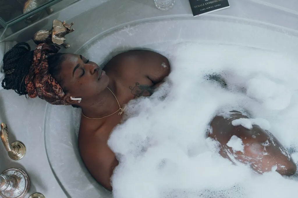 A black queer woman practicing self care and romancing herself in a bubble bath with Airpods on, listening to her favorite podcast.