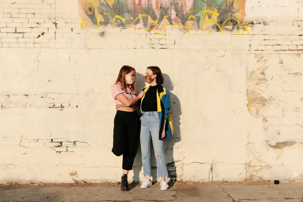 Two queer women in a mono-poly relationship standing before a brick wall with masks on.