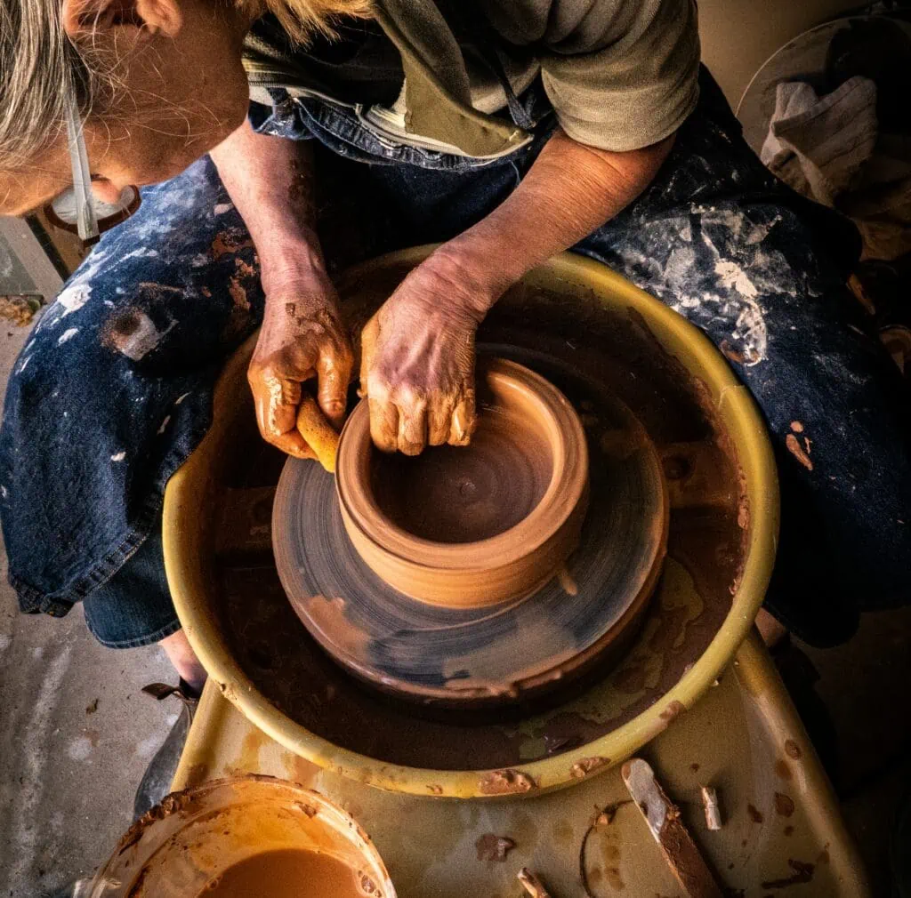 A queer introverted woman in a paint-covered smock shaping a bowl on a ceramics wheel.