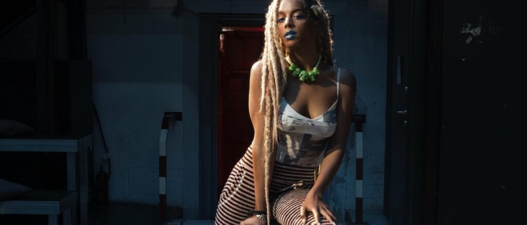 Juliana Huxtable, mujer transexual intersexual negra, posa con leggings a rayas, camiseta blanca de tirantes y largas trenzas decoloradas con un collar de calavera verde al cuello.