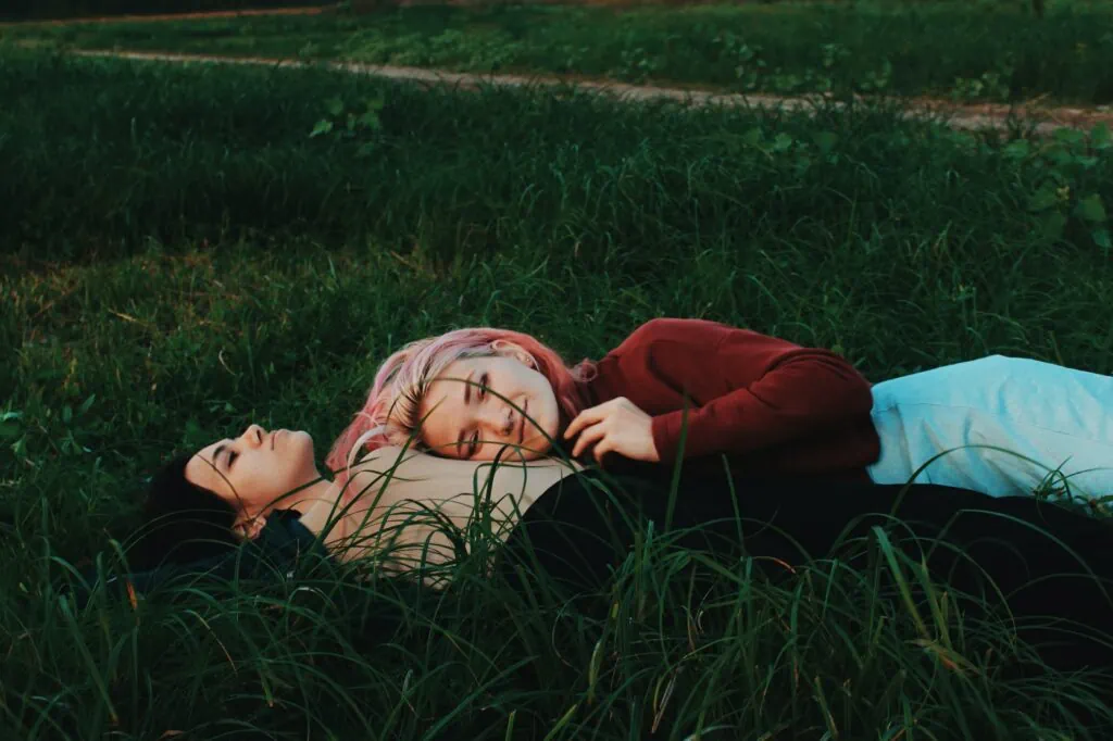 Two lesbian women in a monogamish relationship laying down in the grass together, one in a red sweater with pink hair looks at the camera while laying on the chest of her girlfriend.