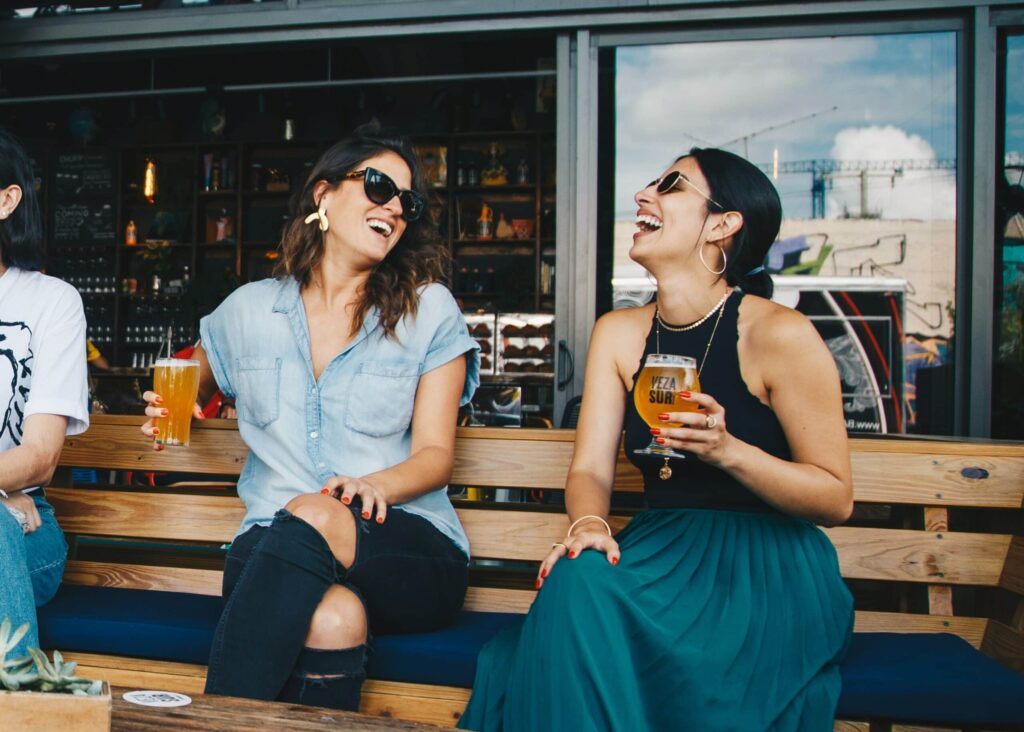Dos mujeres lesbianas sentadas en un banco en una primera cita con cervezas en la mano riéndose de las divertidas preguntas de la primera cita.