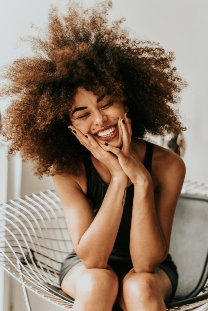 A quirky cute girl with a big afro, crooked teeth, and a mole on her face smiling with her hands on her knees.