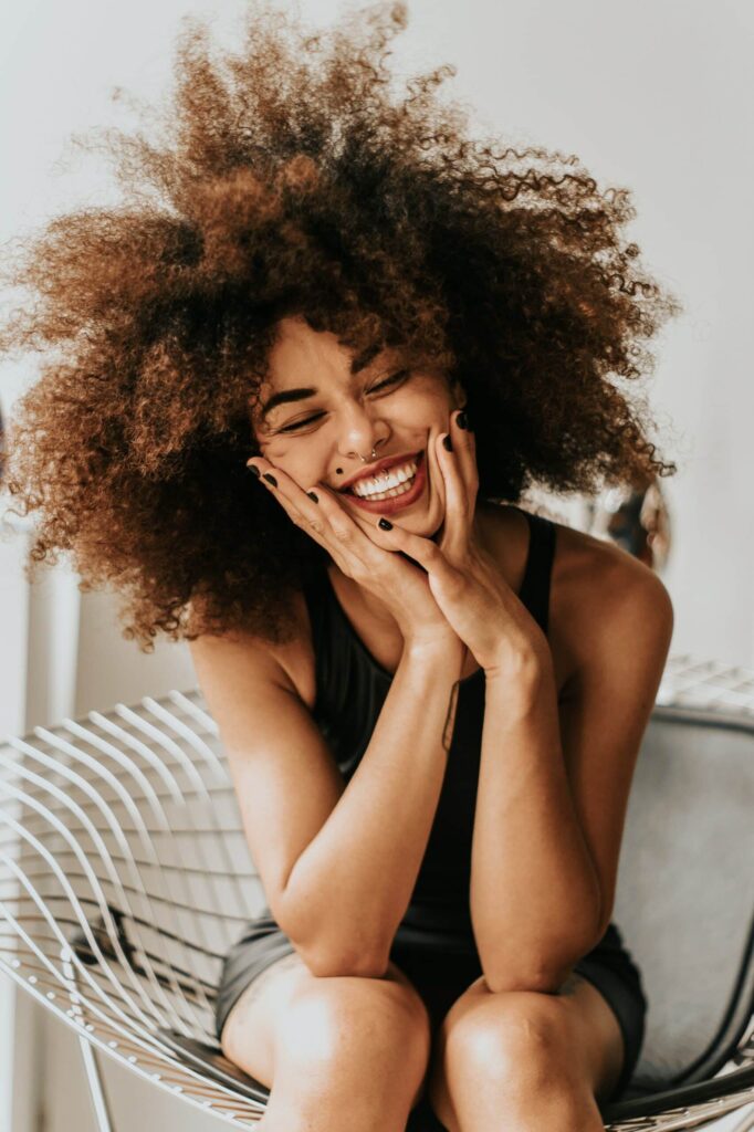 Una chica mona y estrafalaria con un gran afro, dientes torcidos y un lunar en la cara sonriendo con las manos sobre las rodillas.