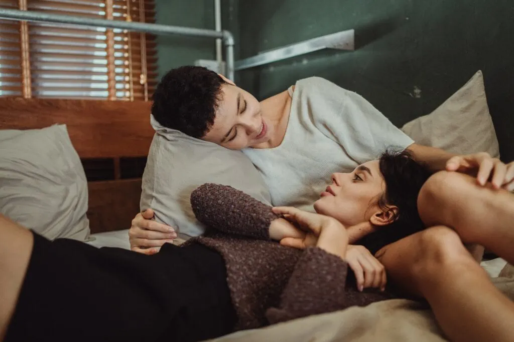  A queer couple on a bed, they’re smiling, talking, and resting on one another.