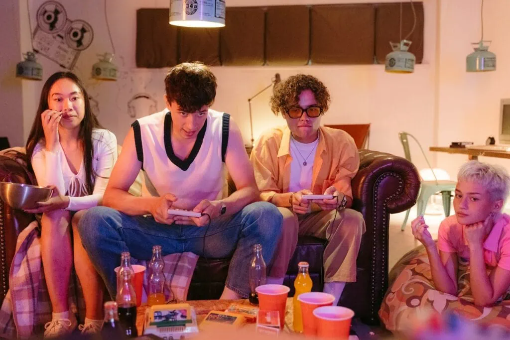 A friend hangout. Two of them are focused on playing a videogame, while the other two are eating snacks while they watch.