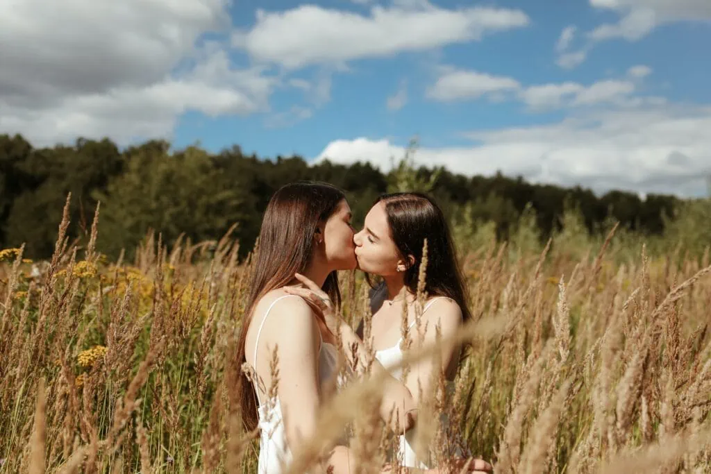 Two lesbian women in a monogamous relationship kissing in a wheat field under a cloudy blue sky.