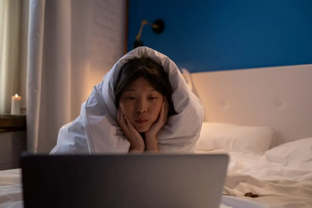 An introverted woman lying in her bed with a duvet pulled over her head in front of a laptop.