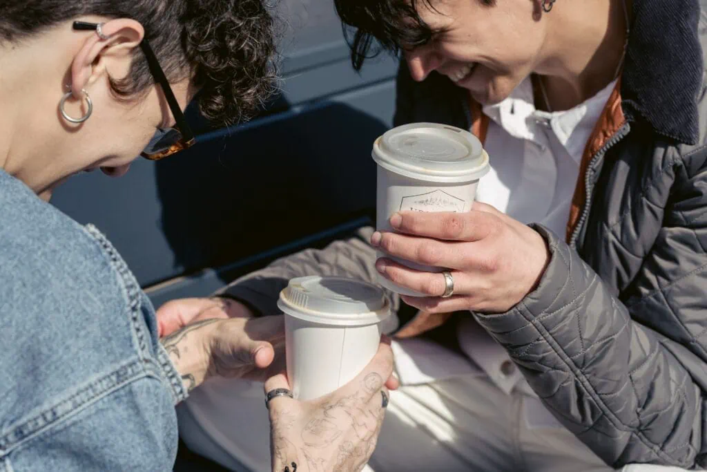 Two androgynous people holding paper coffee cups, and laughing. 