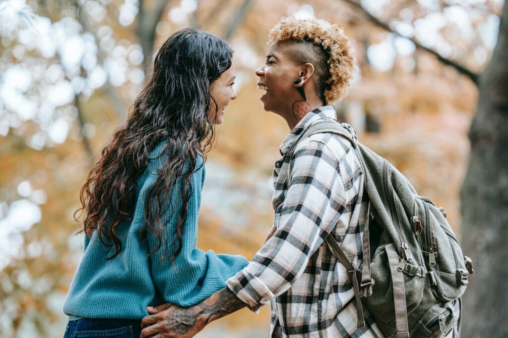 A lesbian couple laughing in the park together after telling some funny first date jokes. One woman is wearing a flannel and the other is in a blue sweater.