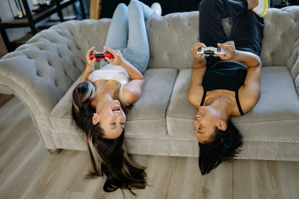 Two women playing a videogame and laughing, both are sitting upside-down on the couch.