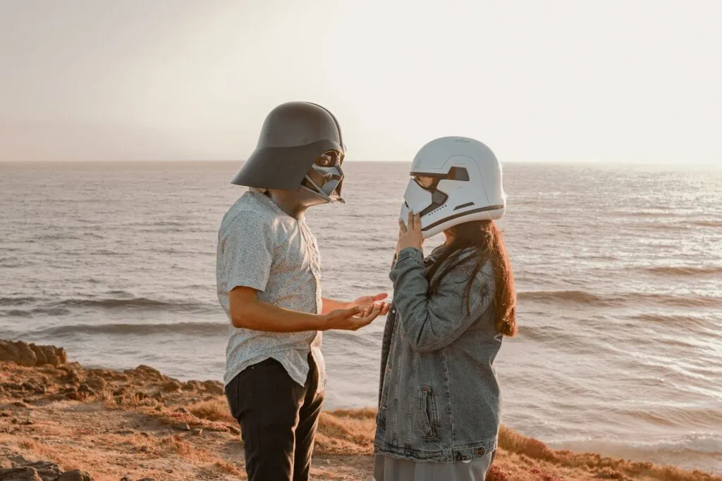 A nerdy couple – one wearing a Stormtrooper helmet, and the other wearing a Darth Vader helmet – face each other on a beach, with the sun setting behind them.