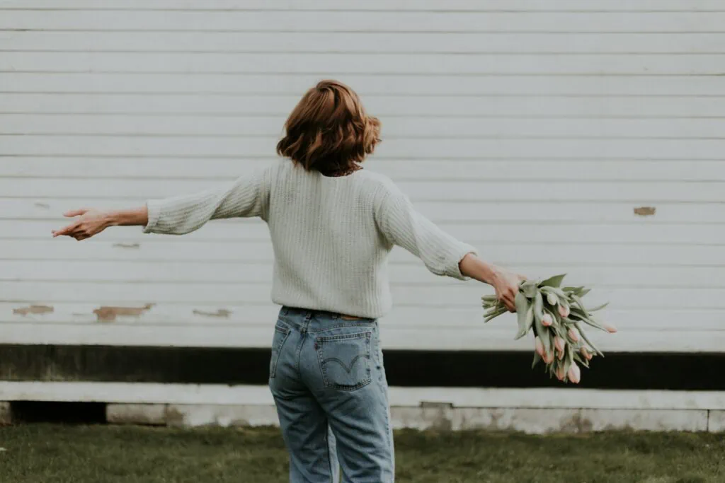 A single woman with her arms outstretched holding flowers in front of a white barn.