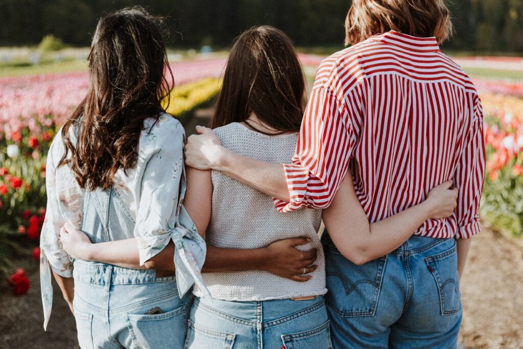 Three queer women in a polyamorous dynamic, with their arms around each other’s backs standing in a garden.