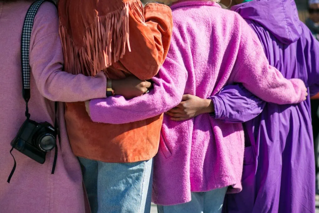 Four polyamorous women in pink, purple, and orange coats with their arms wrapped around each other.