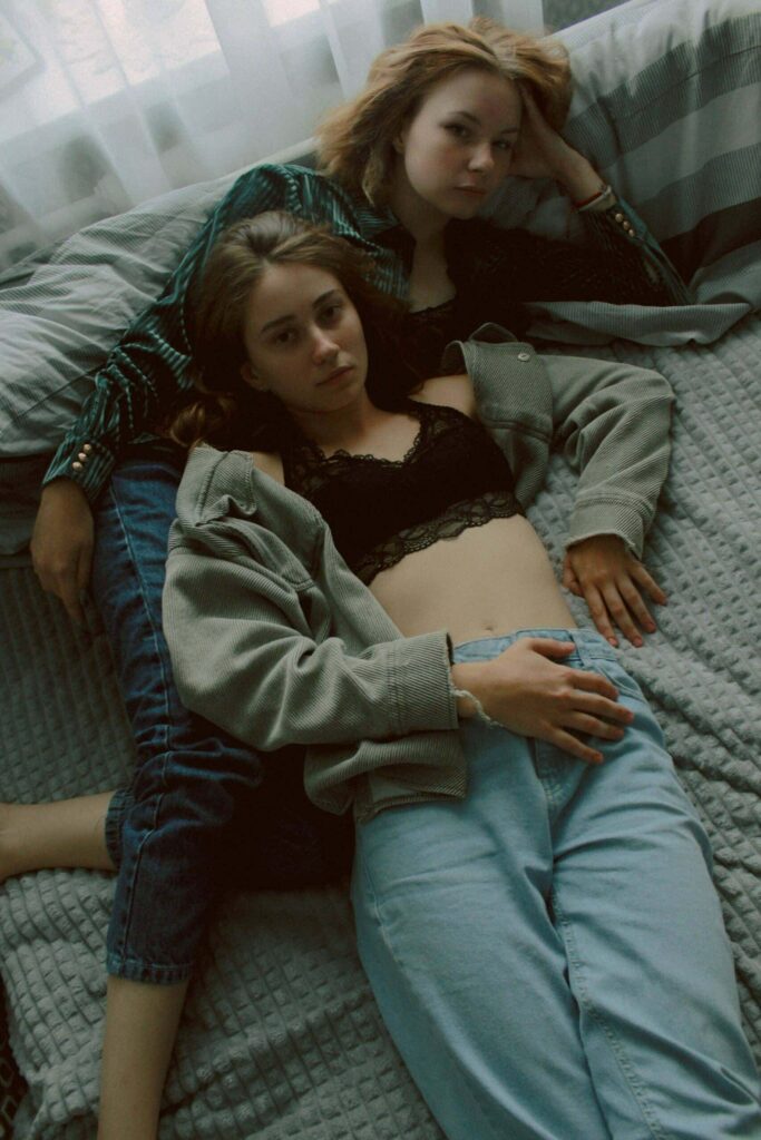Lesbian couple posing in bed, wearing jeans and open flannel shirts while embracing.
