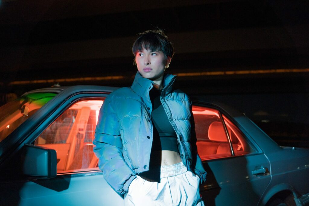 A futch lesbian with a blue puffer jacket and a crop-top standing outside of a sedan late at night.