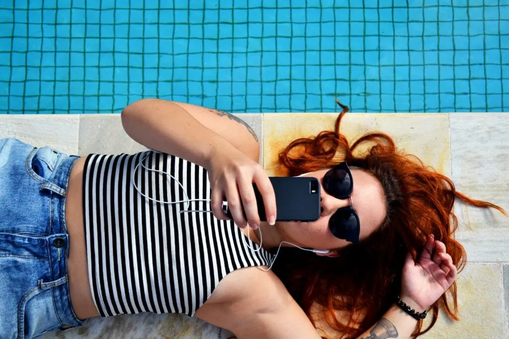 A queer redheaded woman lying by a pool in a striped shirt wearing sunglasses and talking on Facetime.