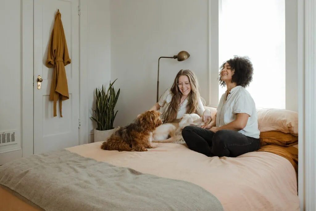Two queer women laughing and sitting on a bed with their dogs after moving in together.