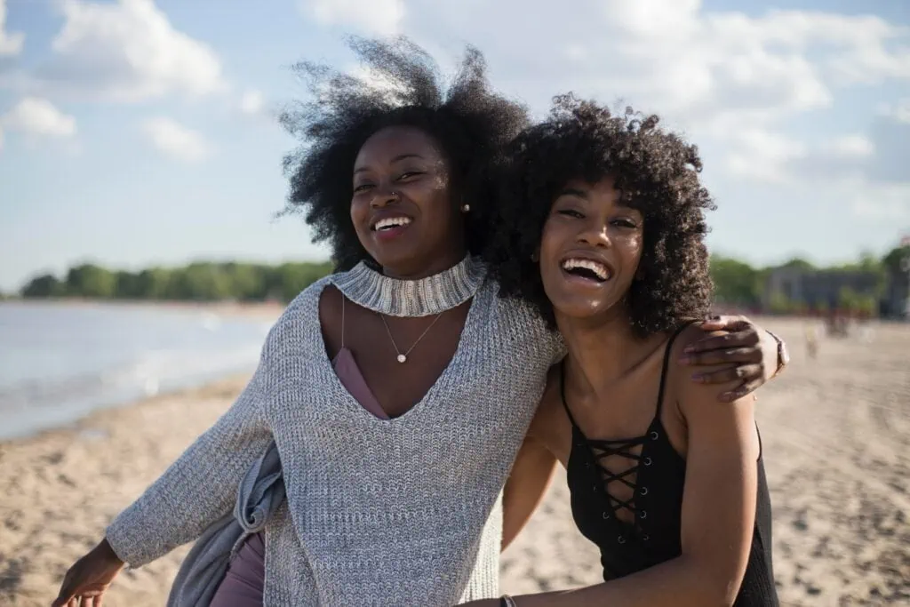 Two black friends laughing and hugging at the beach with the seashore behind them.