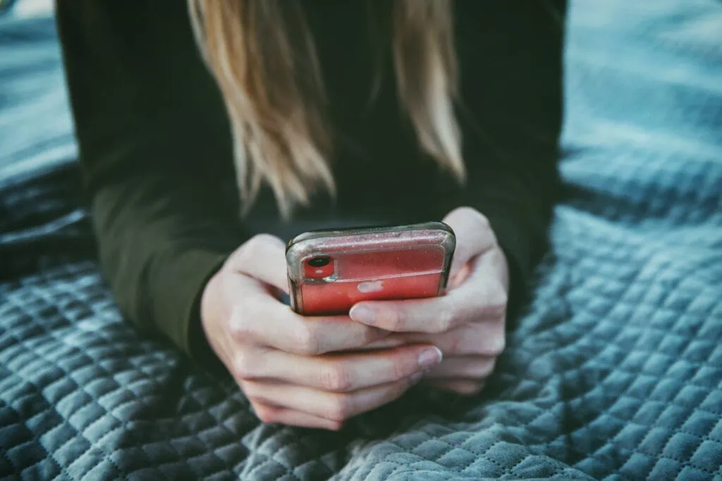 A woman with long blonde hair laying down on a bed on her iPhone, looking for matches on adult dating sites.