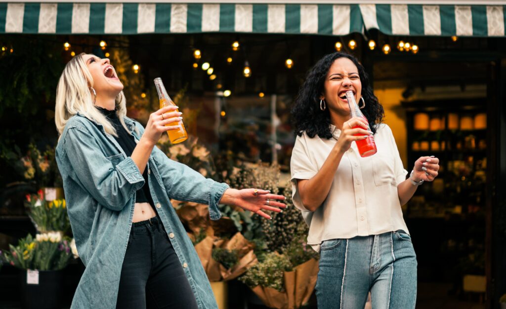 Two queer women in the honeymoon phase of their relationship timeline laughing and drinking Jarritos outside of a shop in New York City.
