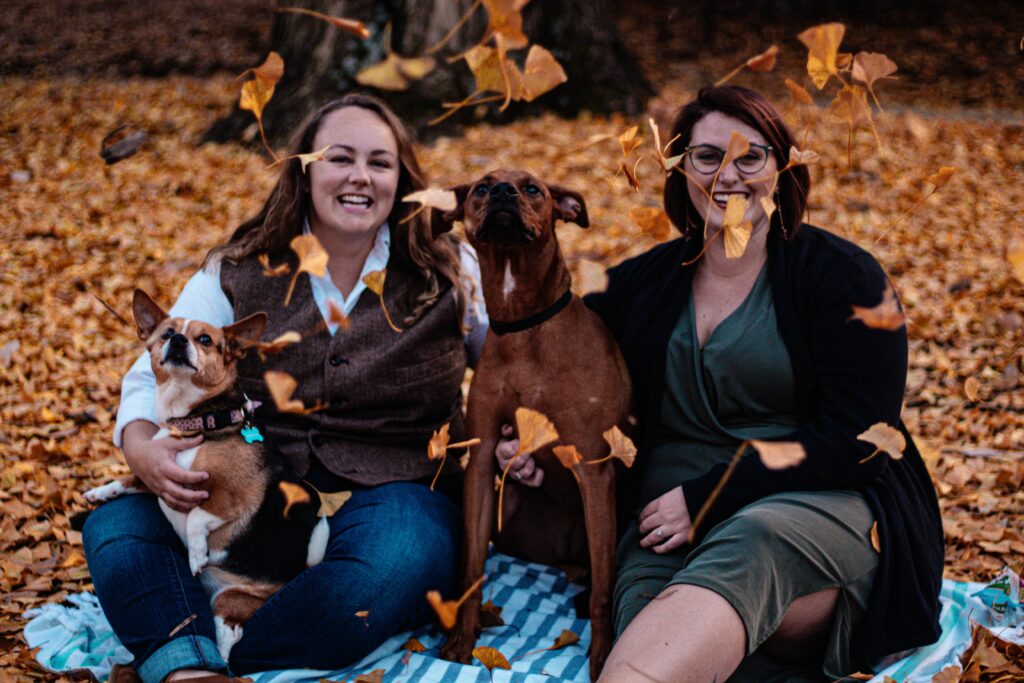 Two lesbian women with their rescue dogs in an autumnal photoshoot with fallen red leaves all around.