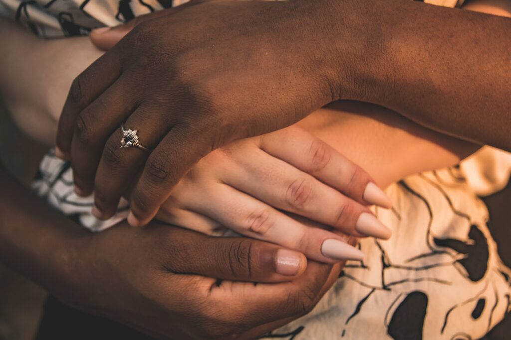 A photo of two interracial sapphic women’s hands intertwined, one with an engagement ring on her signifying the committed phase of a relationship timeline.