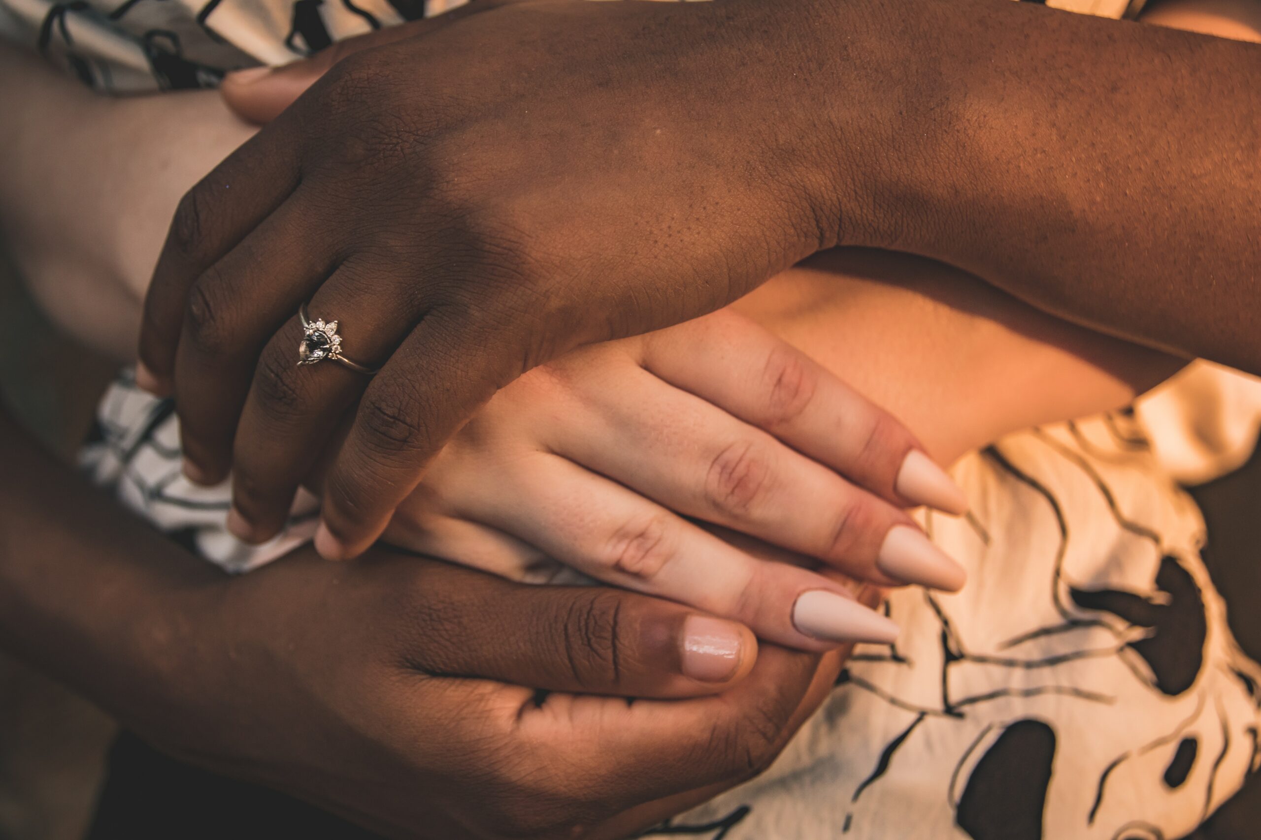 Une photo de deux mains de femmes saphiques interraciales entrelacées, l'une portant une bague de fiançailles signifiant la phase d'engagement d'une ligne du temps de la relation.