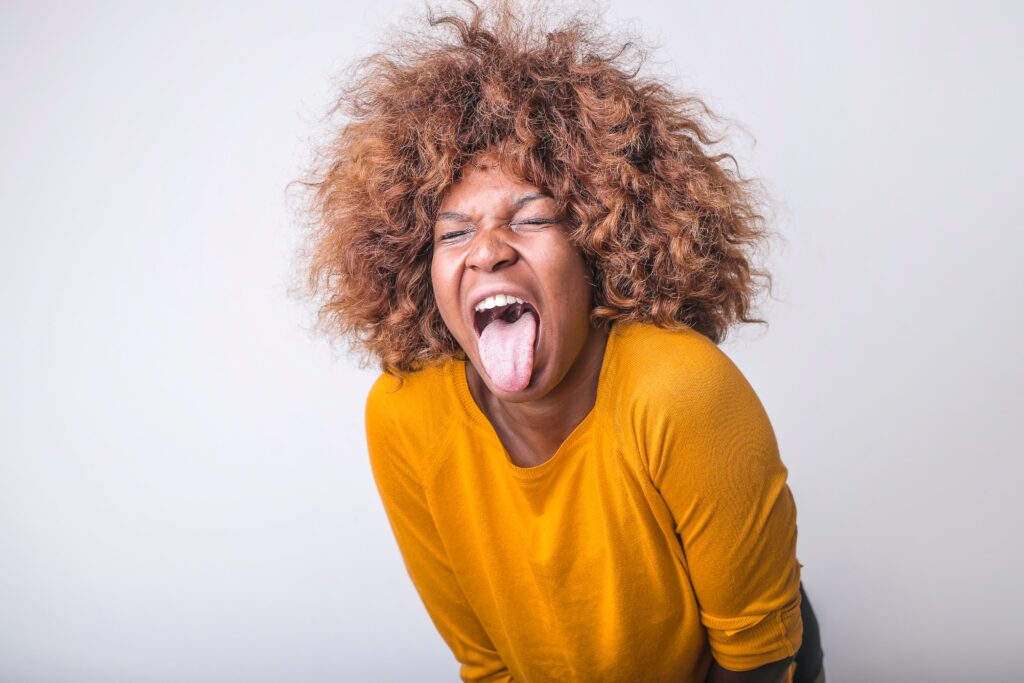 A black woman with radiant copper hair sticking her tongue out in disgust in a yellow long sleeve shirt in front of a light gray background.