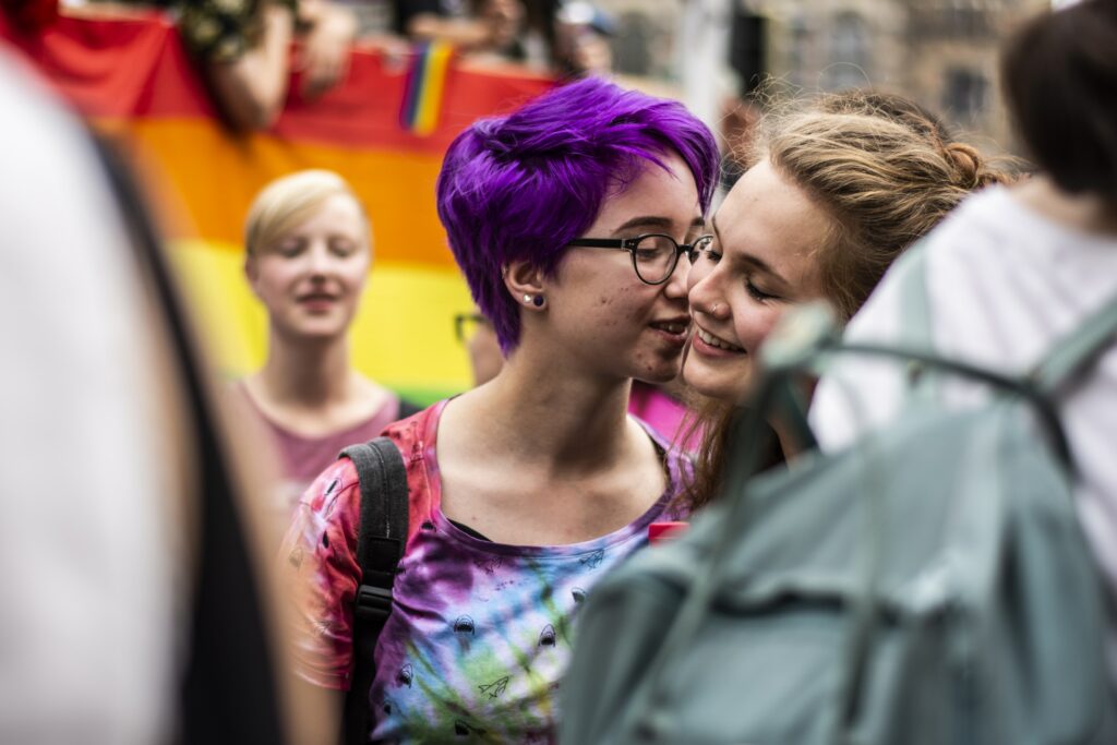 A queer couple embrace as people surround them at a Pride March. A dating goal could be wanting someone to go to a Pride March with.