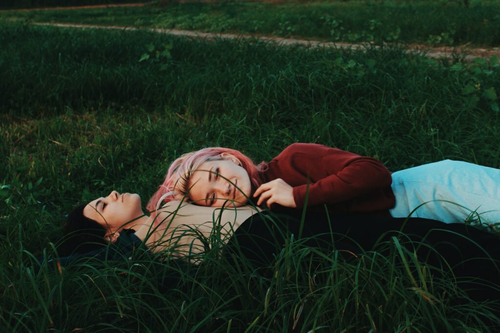 A queer couple lays on the ground, one person on their back and the other resting their head on the other’s stomach. Some would consider this as couple goals.