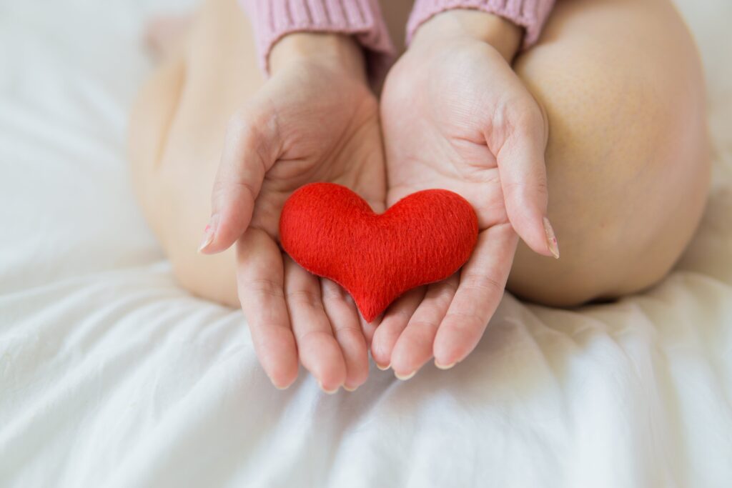 A close-up photo of a woman kneeling on a white bed with her hands outstretched. Inside them is a small felted red heart.