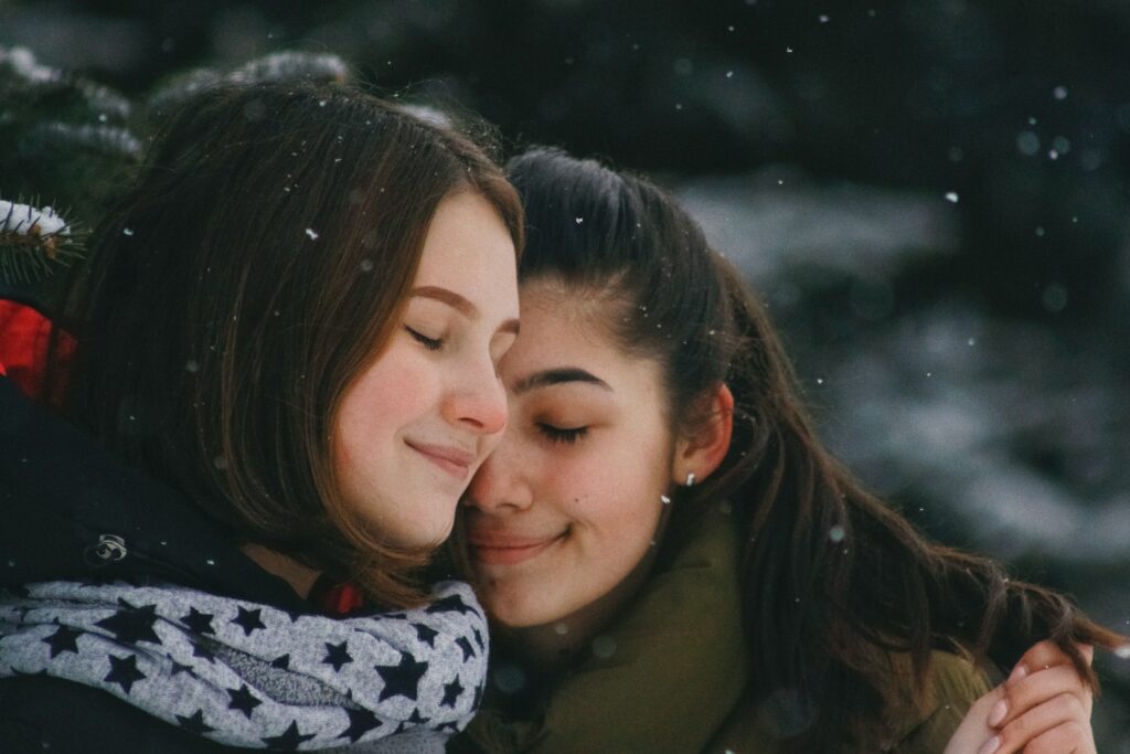 A lesbian couple hug outside while it’s snowing. They’re wearing winter outerwear and look like they have their six human needs met.