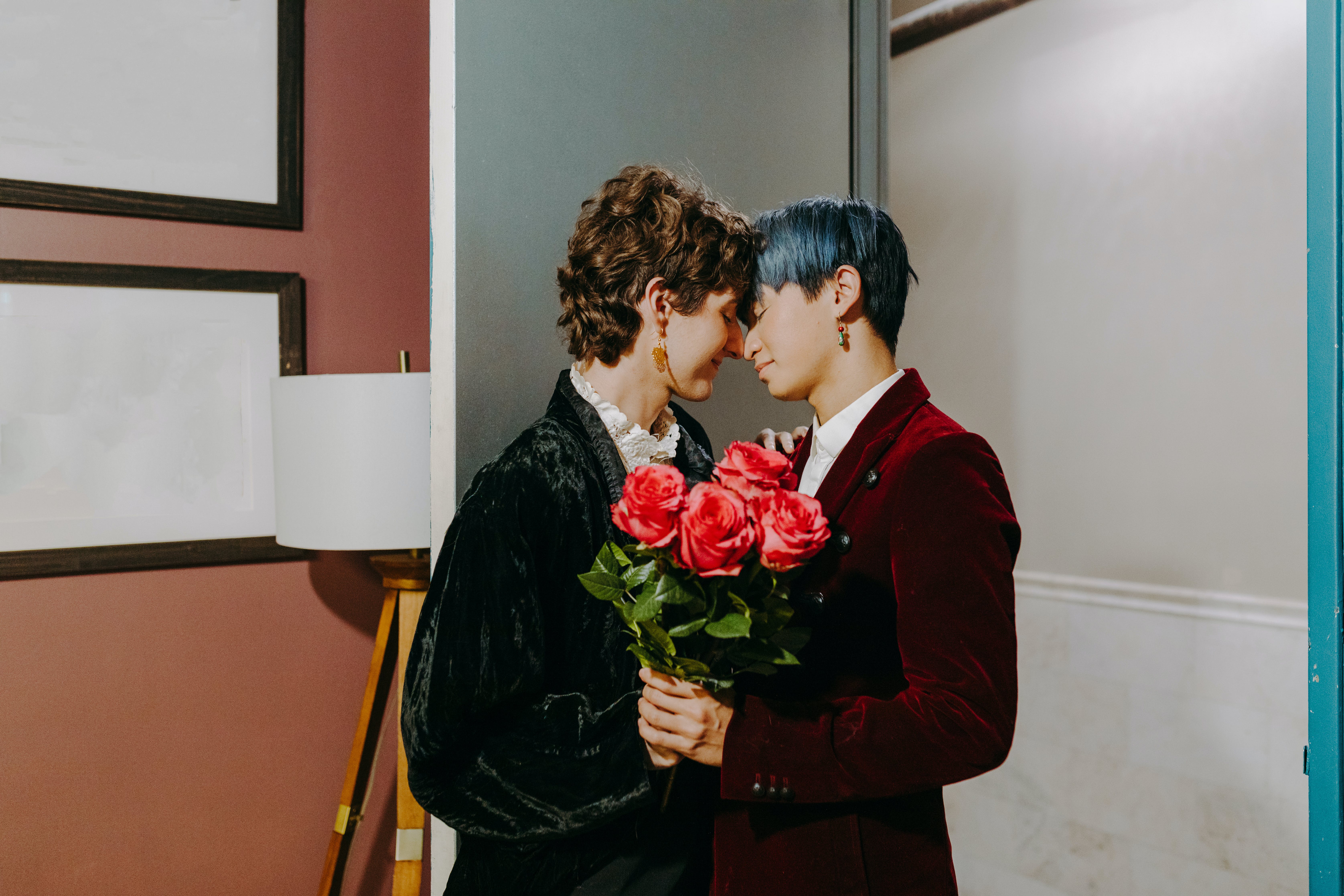 Two young LGBTQIA+ people in a long-term relationship, wholeheartedly in love. They are both dressed in velvet suits with short hair, their faces lovingly pressed together, both holding a bouquet of roses.