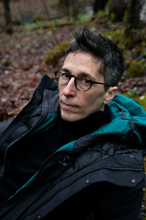 A photo of Alison Bechdel, lesbian cartoonist and graphic comic. She is butch and handsome in round glasses, leaning back in a winter coat with wet moss behind her.