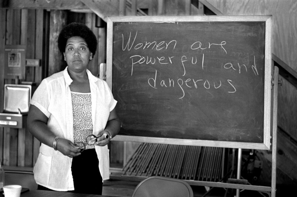 A black and white photo of black lesbian writer and poet Audre Lorde standing in front of a blackboard with writing that reads “Women are powerful and dangerous”
