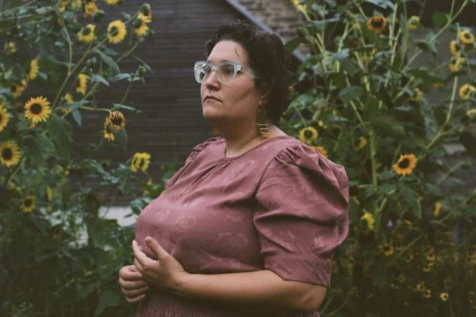 An image of Carmen Maria Machado, queer, polyamorous, and lesbian writer. She is standing in a garden full of sunflowers with a pink dress on.