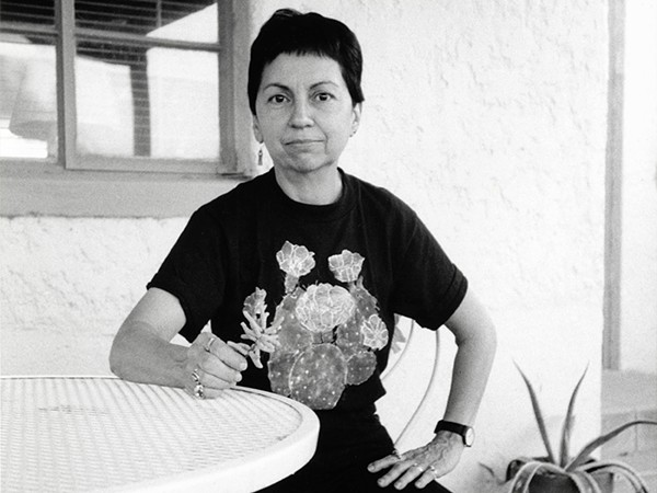 A black and white photo of Gloria Anzaldúa, Chicana feminist and lesbian writer, sitting at a table with short black hair. Her hand is resting on her leg and she is looking at the camera.