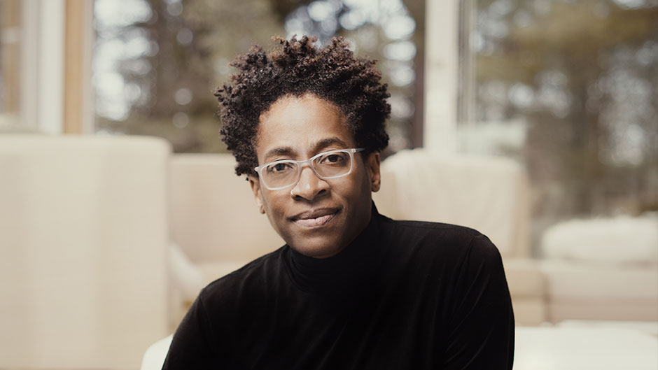 An author portrait of Jacqueline Woodsoon, black lesbian writer of color, in a black turtleneck and white glasses looking boldly at the camera.
