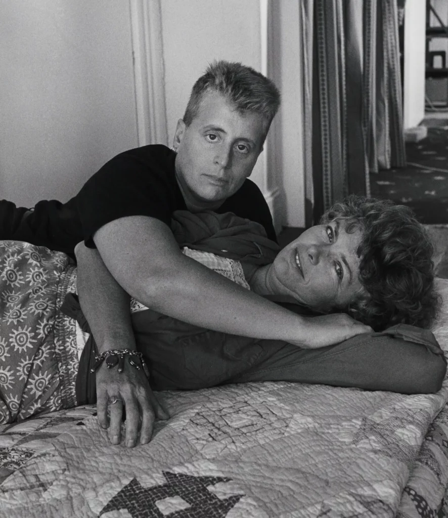 An iconic black and white photo of trans lesbian writer Leslie Feinberg and hir partner Minnie Bruce Pratt lying on a bed together, both looking at the camera.