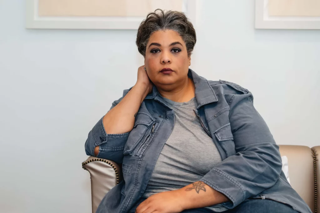 Author portrait of Roxane Gay, lesbian essayist and memoir writer. She is sitting in on a cream-colored couch in a blue lightweight jacket looking slightly past the camera