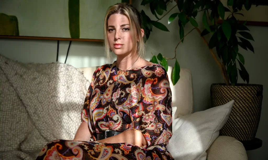 A striking image of trans lesbian author Torey Peters at her home in Brooklyn. She is wearing a multi-colored dress on a beige couch with a plant blooming behind her.
