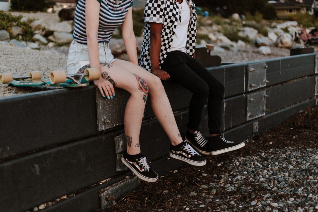 Two cute queers sitting on a ledge with matching black and white striped and checkered outfits on, with a skateboard by their side. This could be you meeting up with the girl of your dreams after you’ve nailed your dating chat on the apps.