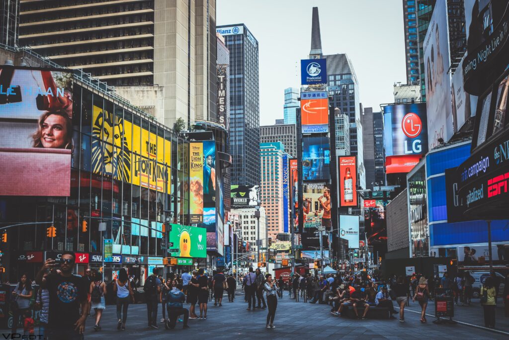 A vibrant and bustling scene of Times Square in New York City, capturing the essence of the city's energy and diversity. The landscape features towering digital billboards displaying colorful advertisements, crowds of people walking along the pedestrian-friendly streets, iconic yellow taxis, and the famous red steps in the background, under a clear blue sky.