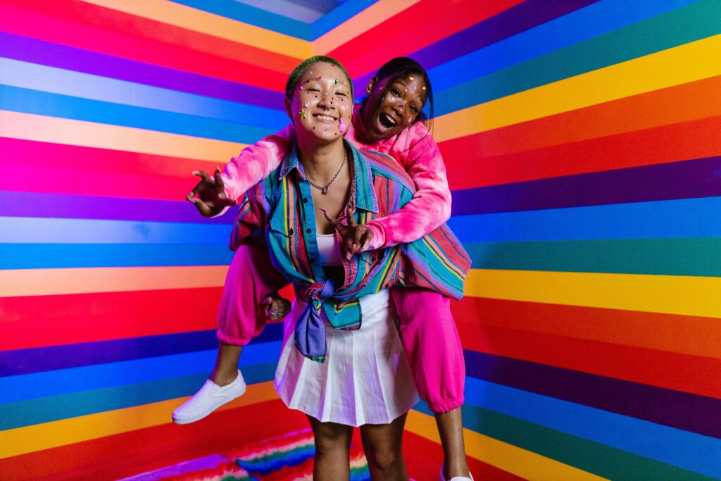 Two queer women joyfully playing in a gym or wellness space, with an LGBT flag in the background. They are smiling and appear to be having a great time, surrounded by equipment indicative of a fitness environment in New York. The atmosphere is welcoming and inclusive, emphasizing the celebration of LGBT identities.