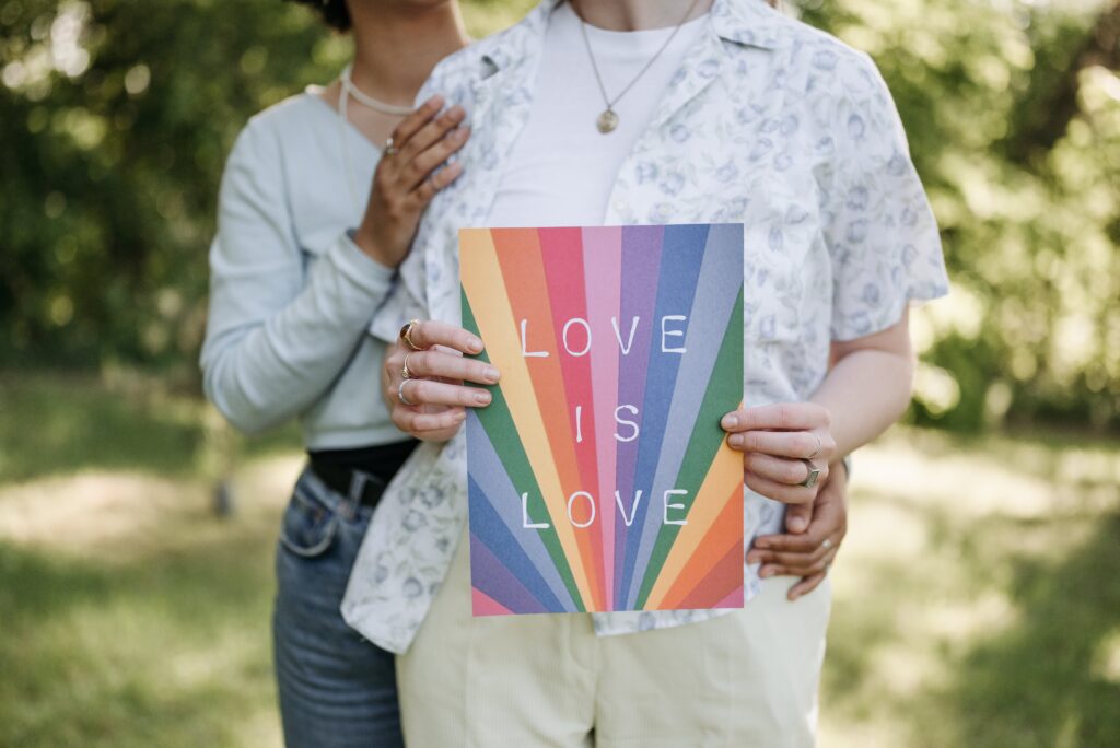 Two LGBT women holding a poster that reads 'Love is Love', expressing unity and support for the LGBT community. A vibrant hub for health and human rights advocacy, with LGBT-themed posters and rainbow flags, symbolizing a safe and inclusive environment.