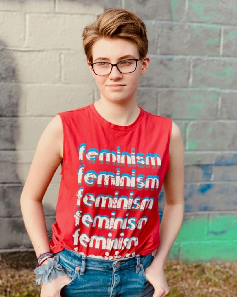 An empowered queer woman poses confidently in front of an artistically decorated wall. She sports a bold haircut from Atelier Upton, complementing her striking appearance. Her attire features a prominent 'feminism' shirt, symbolizing her advocacy and beliefs.