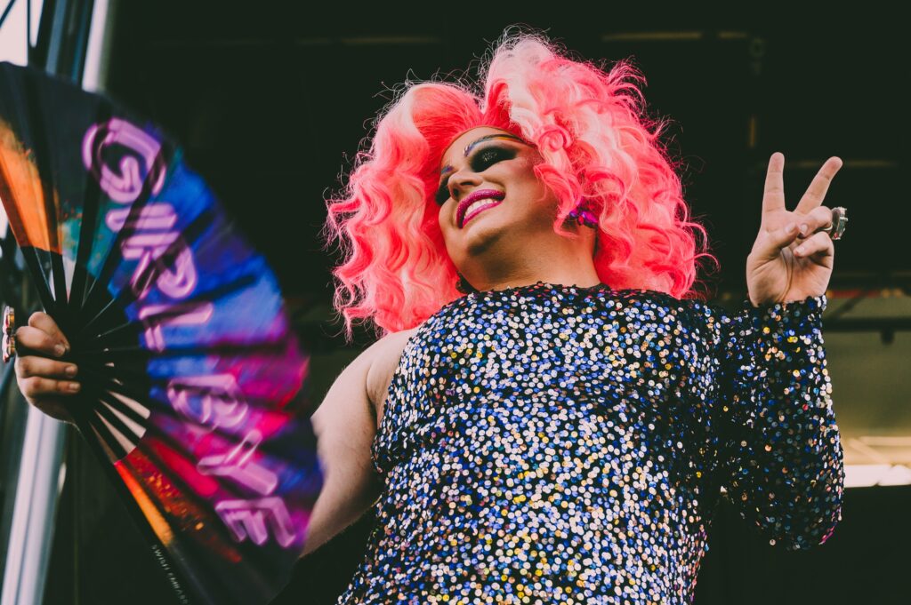A vibrant image of a drag queen, exuding confidence and flair, as she waves a fan emblazoned with the words 