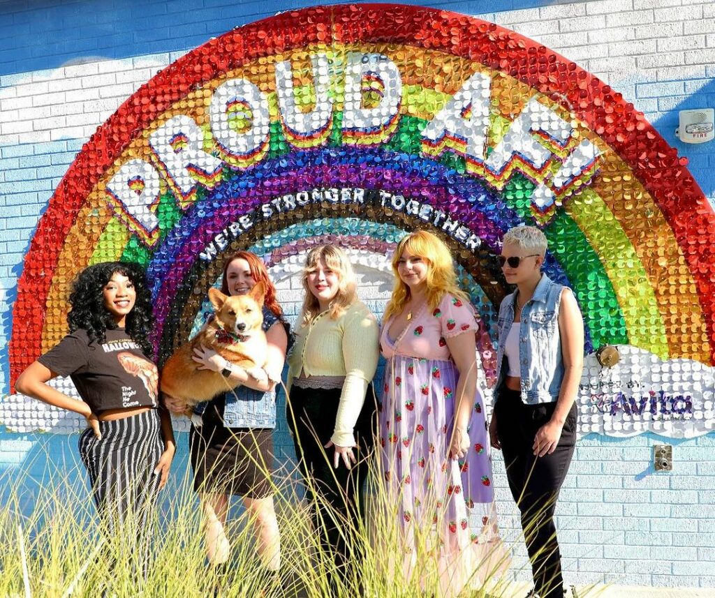 An enthusiastic group of LGBTQ+ individuals from the Beauty Parlour salon, proudly standing together in front of a sign that reads 'Proud AF! We are stronger together', showcasing their solidarity and support for the queer community.